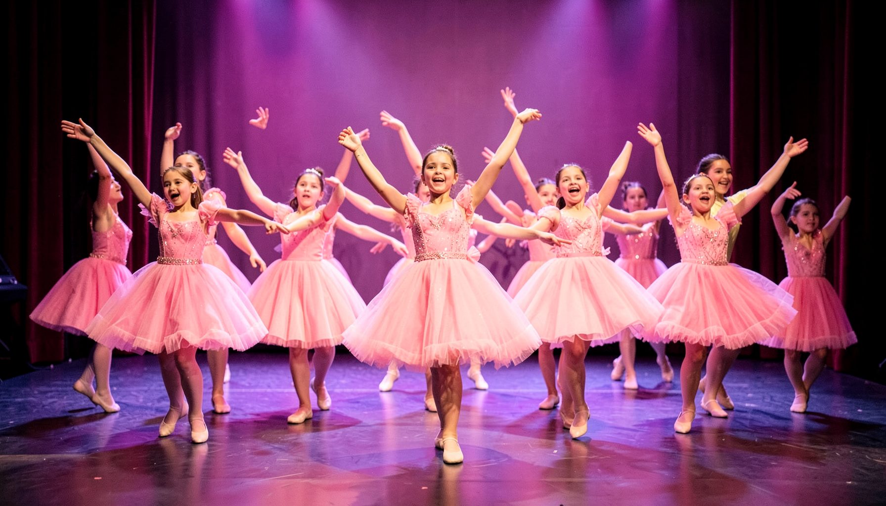 Grupo de niñas con uniforme de danza color rosa realizando una coreografía sincronizada en un escenario iluminado